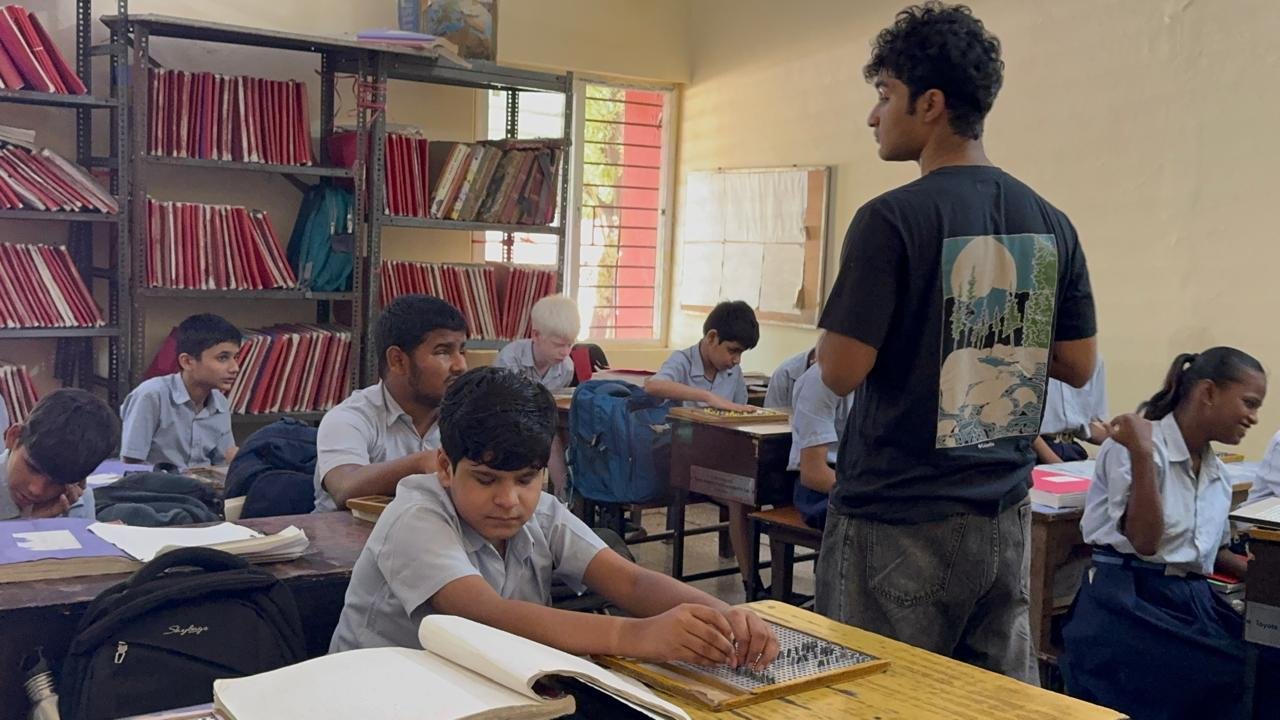 Addressing the class from the front of the room, with a student practicing on a braille slate