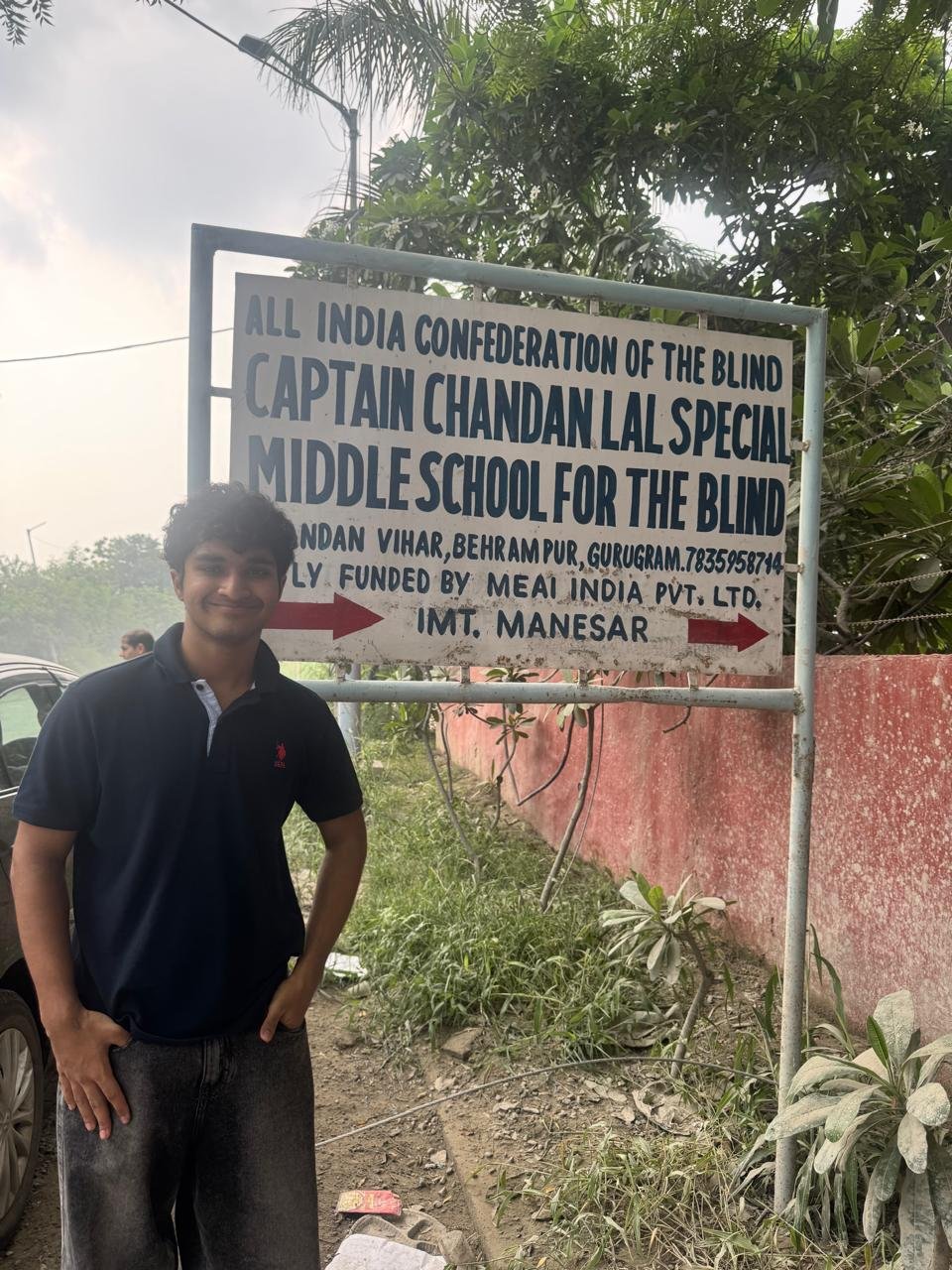 Standing beside the sign for the All India Confederation of the Blind, Captain Chandan Lal Special Middle School for the Blind