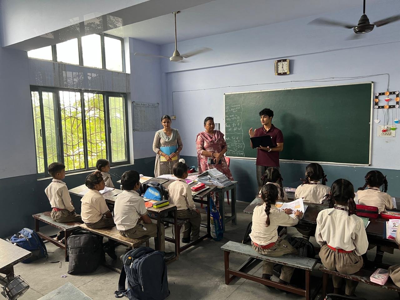 Addressing the classroom with teachers standing nearby and students seated at their desks